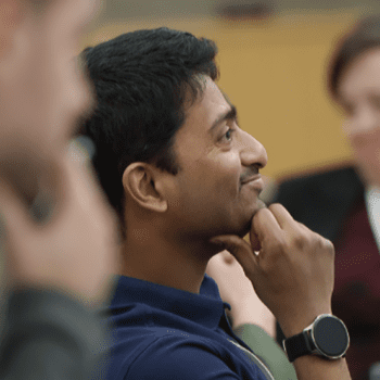 Photo of Students in a classroom. The student in the background is blurred but looks to be a female. The two students in the foreground are male, both looking either at the professor or the board, focused with hand resting on chin.