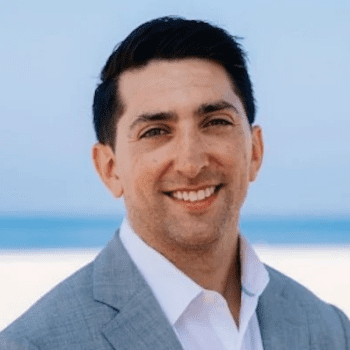 Photo of Chris Murray, MBA 25. White male, wearing a white collar top, blue blazer. Smiling against natural sand and beach backdrop.