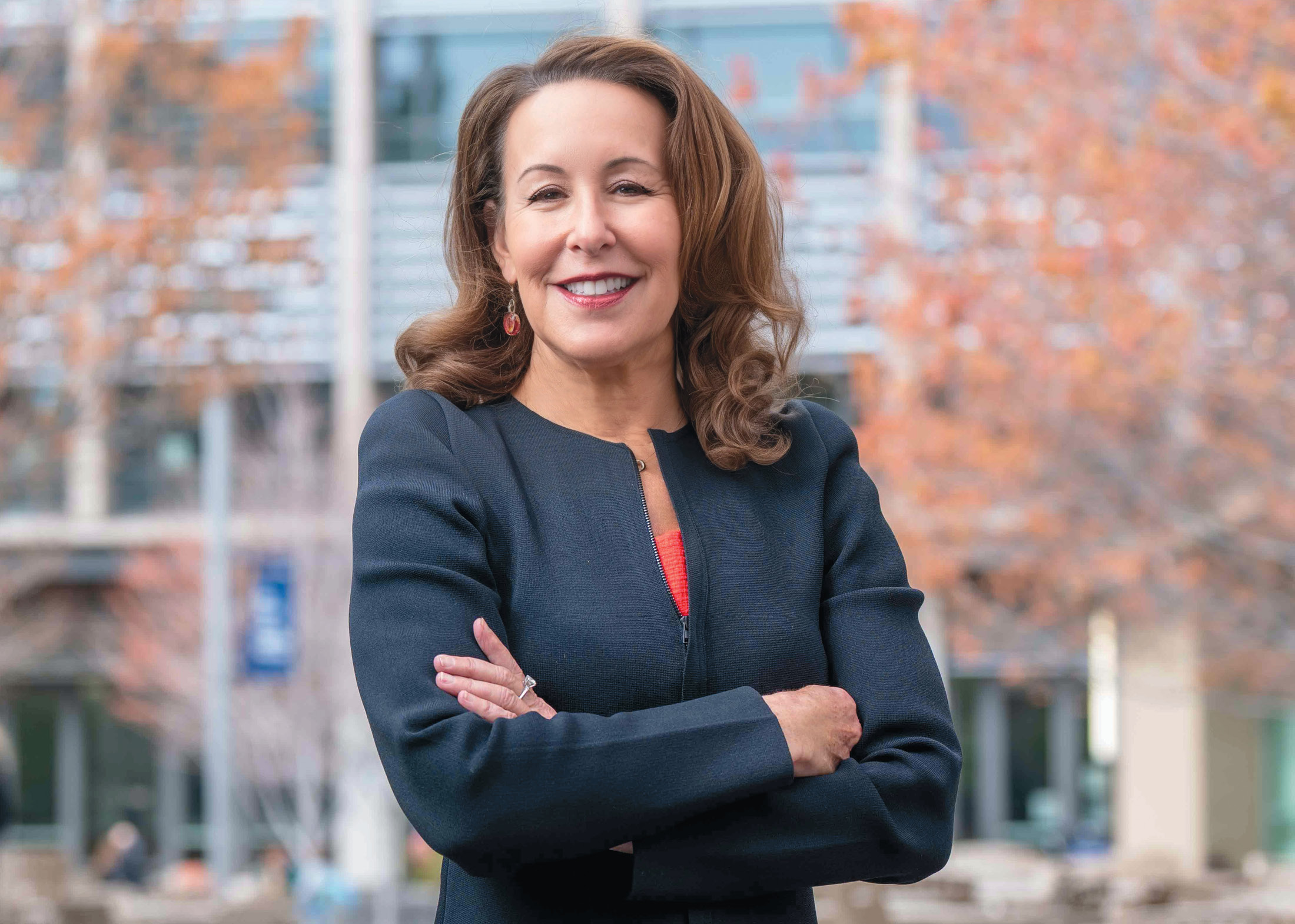 Smiling person in blue jacket stands in front of a building with arms crossed