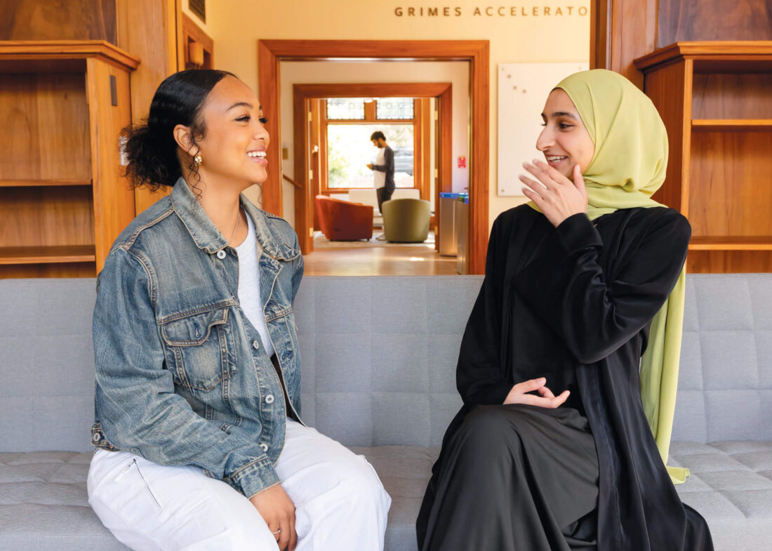 A student in a denim jacket and white pants sits on a grey couch talking to a person in a black dress and light green hijab.