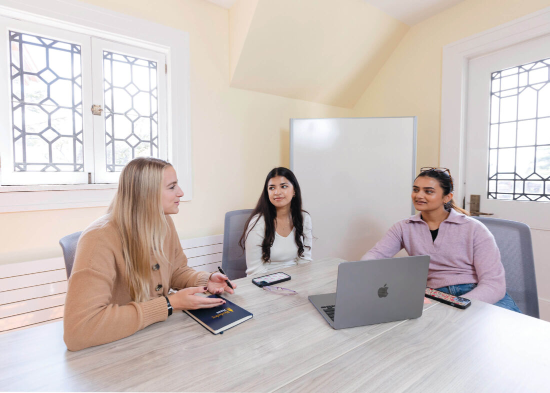 Three people sit at a light colored table containing a notebook, cell phones and a laptop