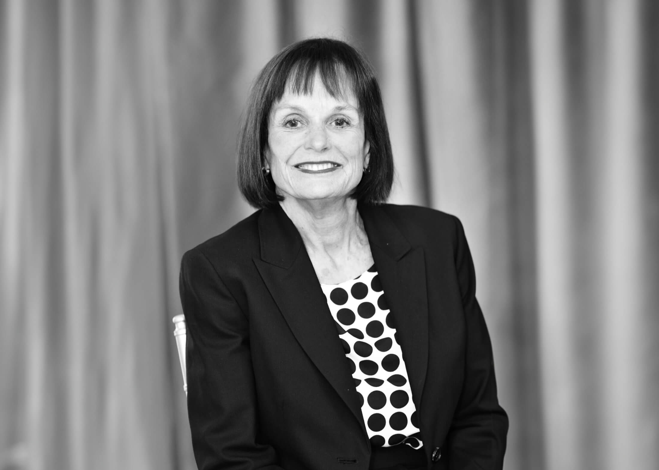 Person in black blazer over a white shirt with black dots on it sits in a chair in front of a backdrop