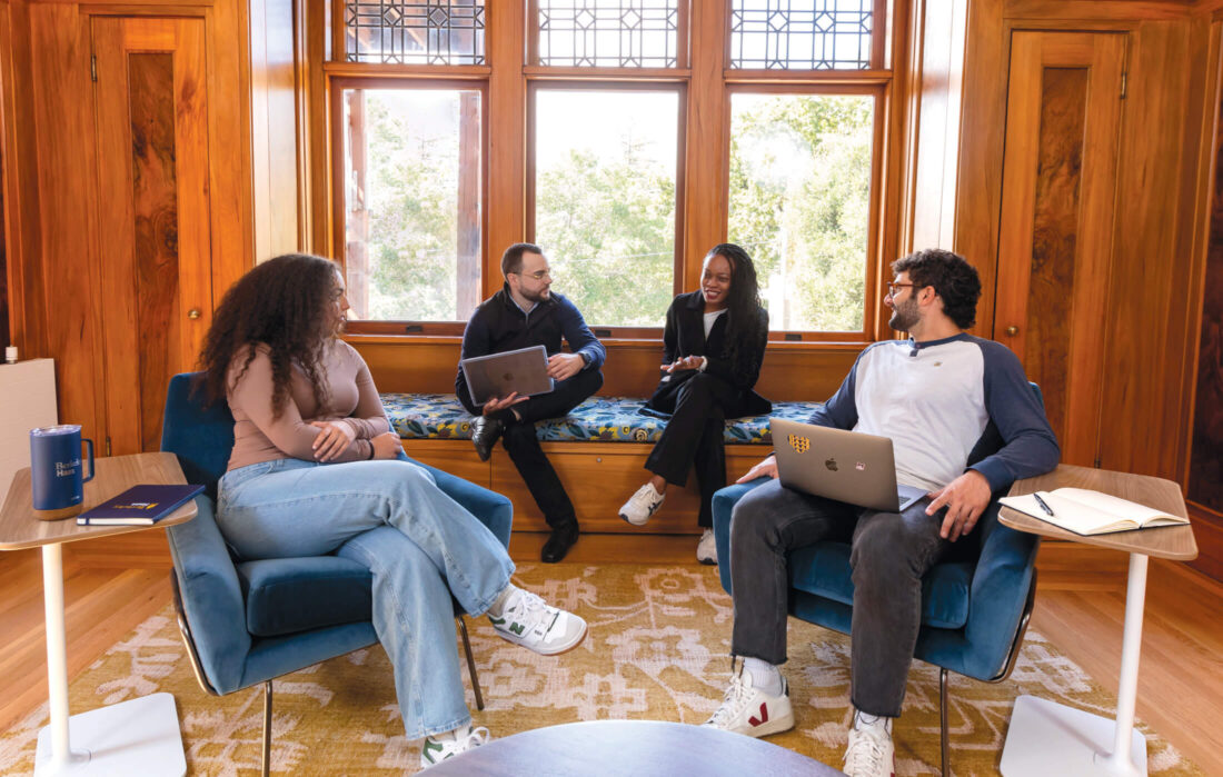 Two students sit in blue arm chairs, looking over their shoulders at two others sitting on a window seat. 