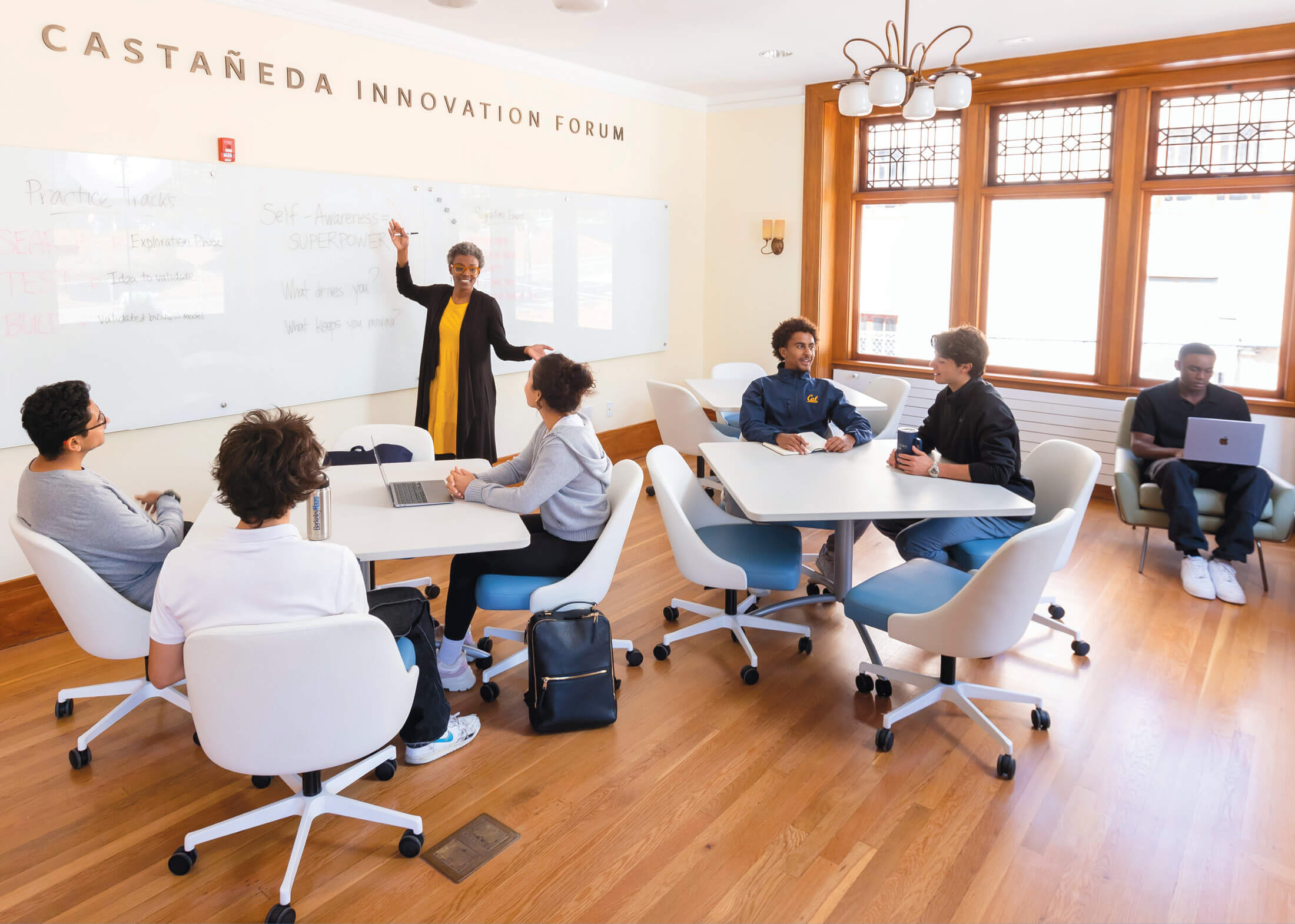 Students seated in white rolling office chairs at white square tables while a person stands in front of a white board on one side of the room