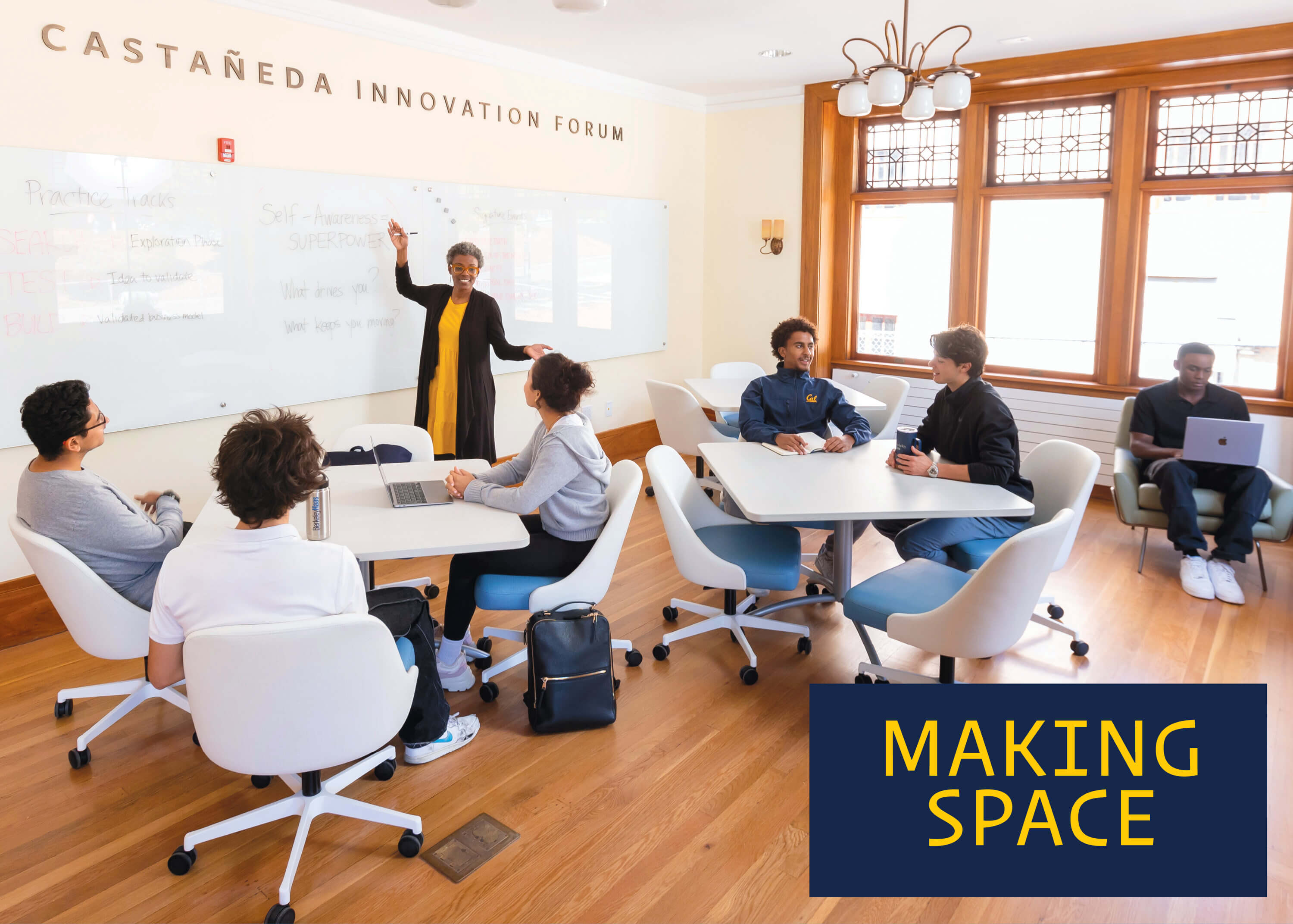 Students seated in white rolling office chairs at white square tables while a person stands in front of a white board on one side of the room