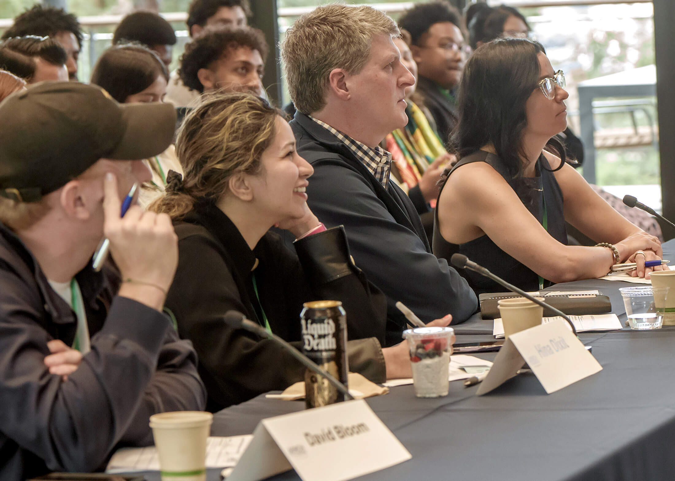 Four people in black clothing sit at a table with microphones and place cards displaying their names while other people are seated in rows behind them.