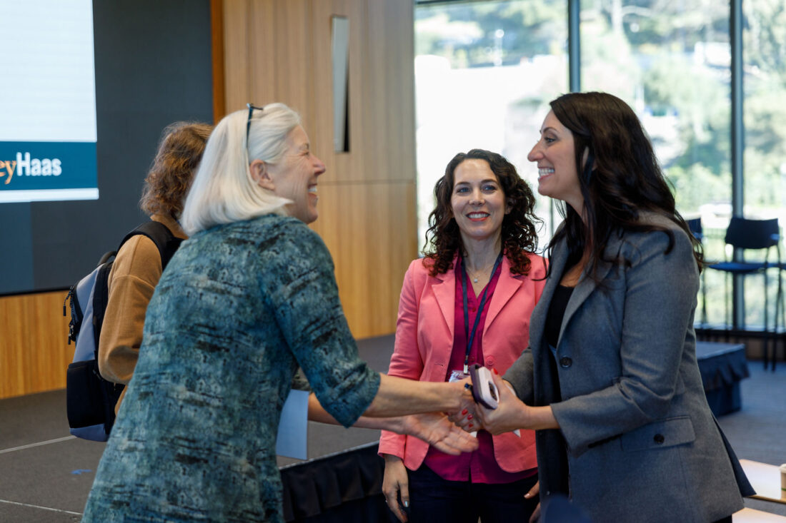 Two women shake hands and smile while another watches and smiles.