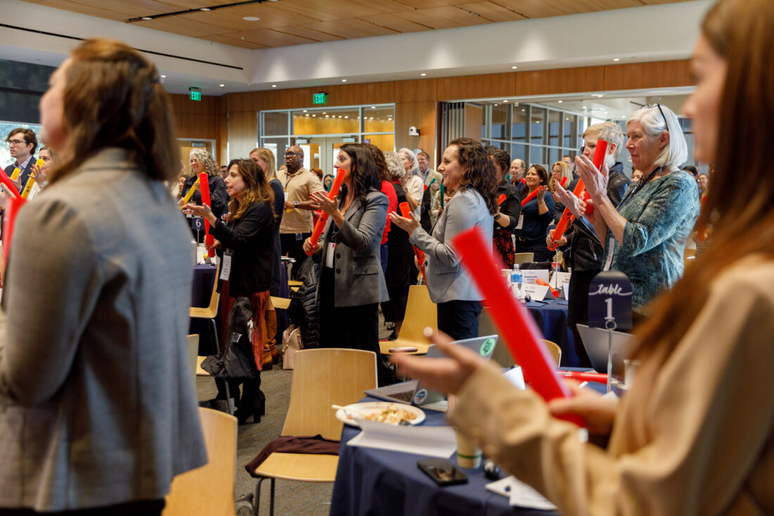 A crowd of people hold orange plastic tubes that they hit against their hands to produce a note.