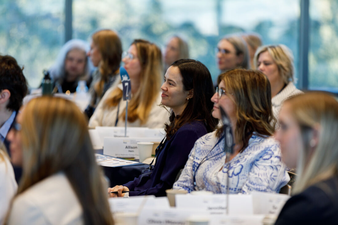 Crowd shot focused on a young woman sitting at a table and listening to a speaker.