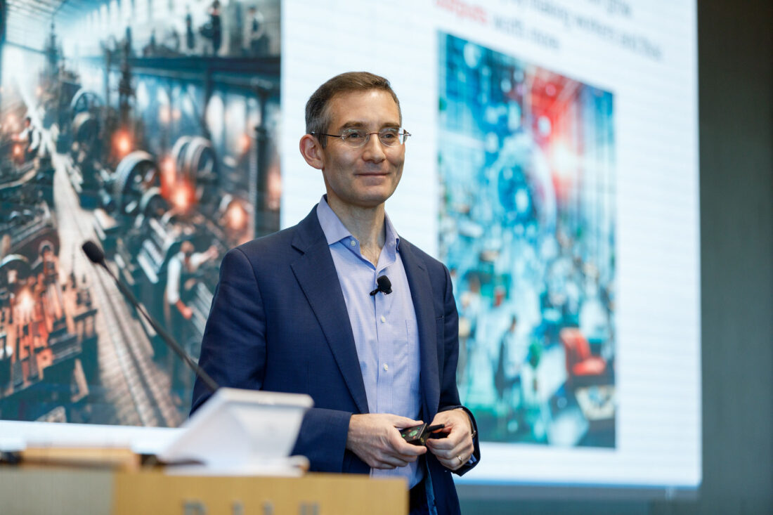 A man wearing glasses and a suit stands at a podium, smiling, with colorful images on a screen behind him