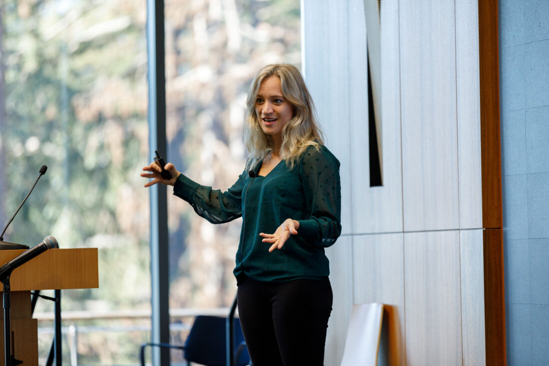 A woman with long blond hair and forest green blouse stands on a stage speaking