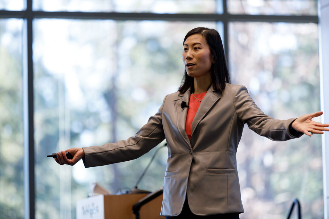 A women in a tan suit jacket and red shirt stands on stake, speaking with her arms opened to the side.