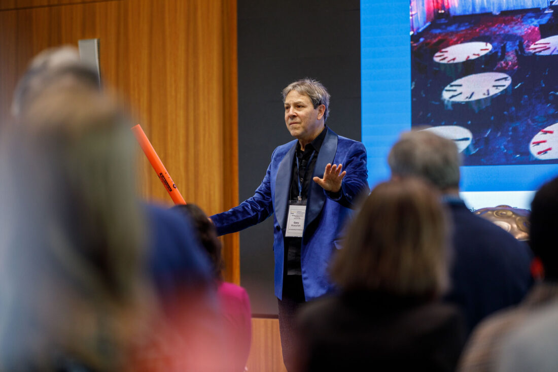 Shot from the perspective of the crowd, this photo shows a man onstage in a purple tuxedo jacket holding an orange plastic tube and directing the crowd with his hands.