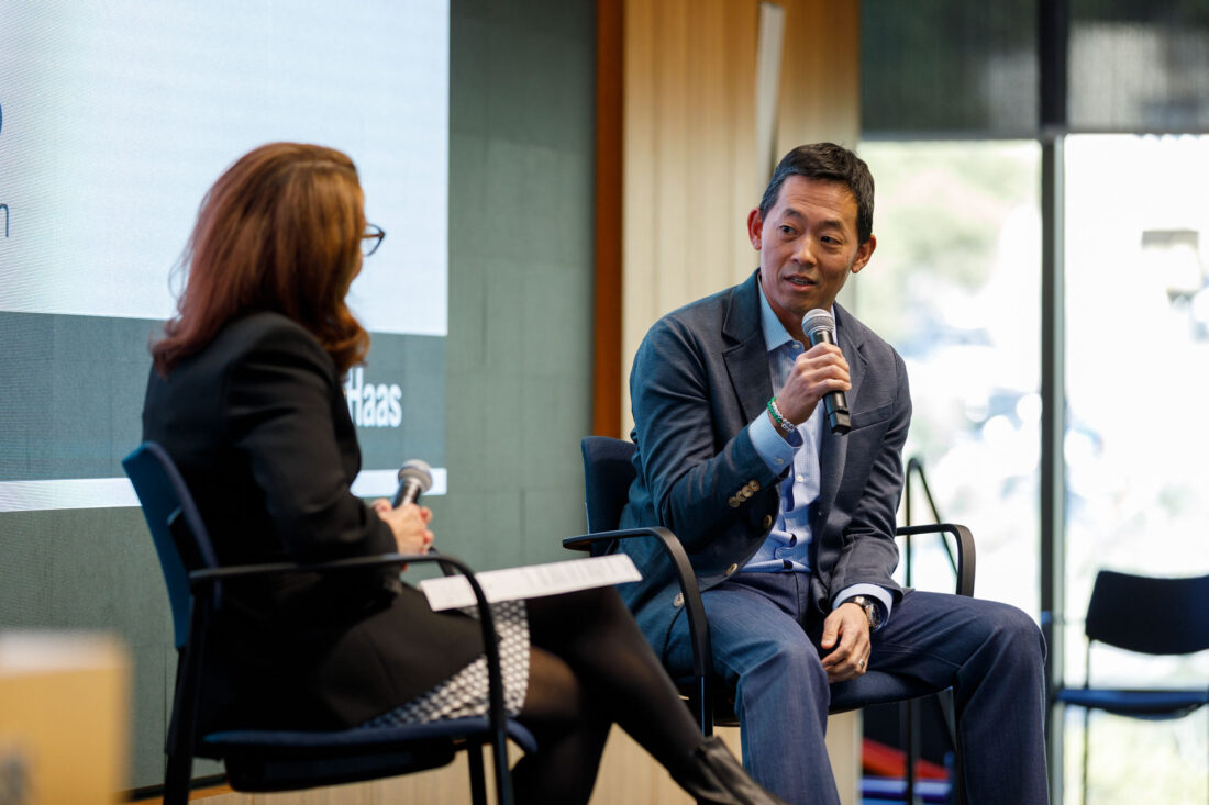 A woman listens to a man in a suit speaking into a microphone. The two are seated onstage.