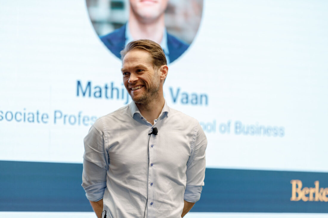 A man in a white dress shirt an lavalier mic stands on a stage, smiling and looking off to the left.