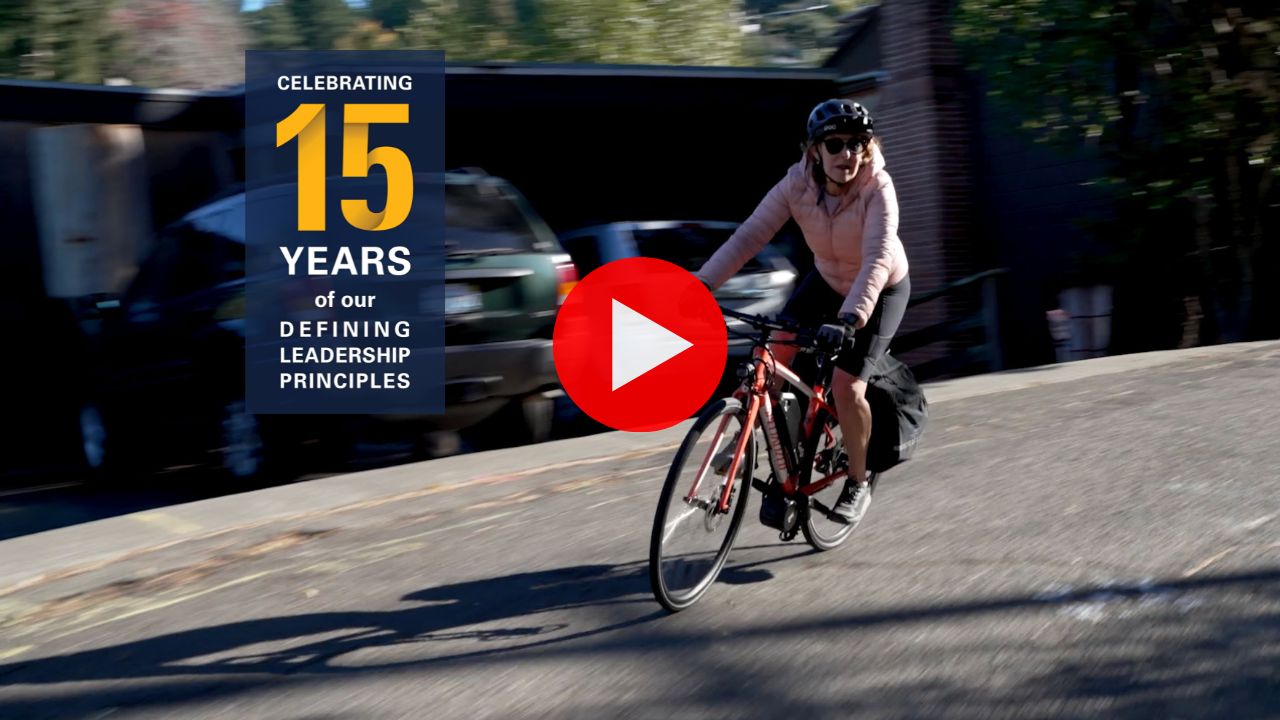 A screenshot of a YouTube video shows a woman in a pink coat biking down a hill. A logo on the screen says "Celebrating 15 years of our Defining Leadership Principles."