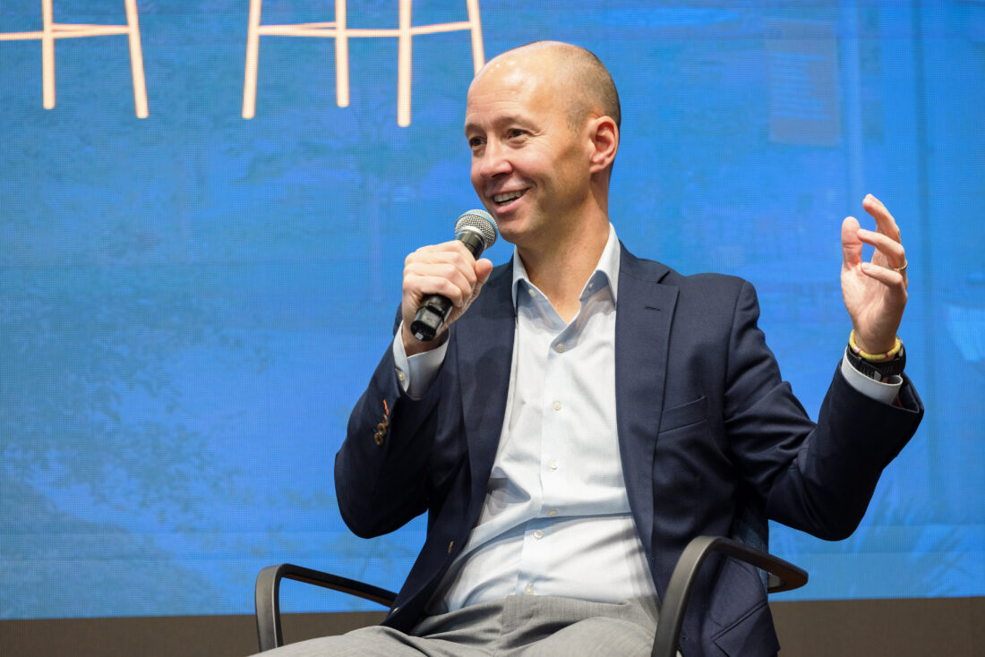 A man with bald head, dress shirt, and suit jacket sits on stage and speaks into a microphone while gesturing with his left hand.